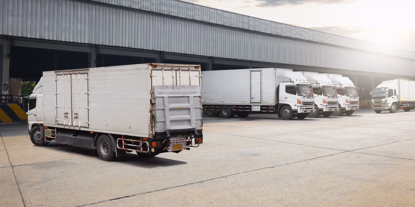 Several white cargo trucks parked outside a large warehouse with a concrete loading area.
