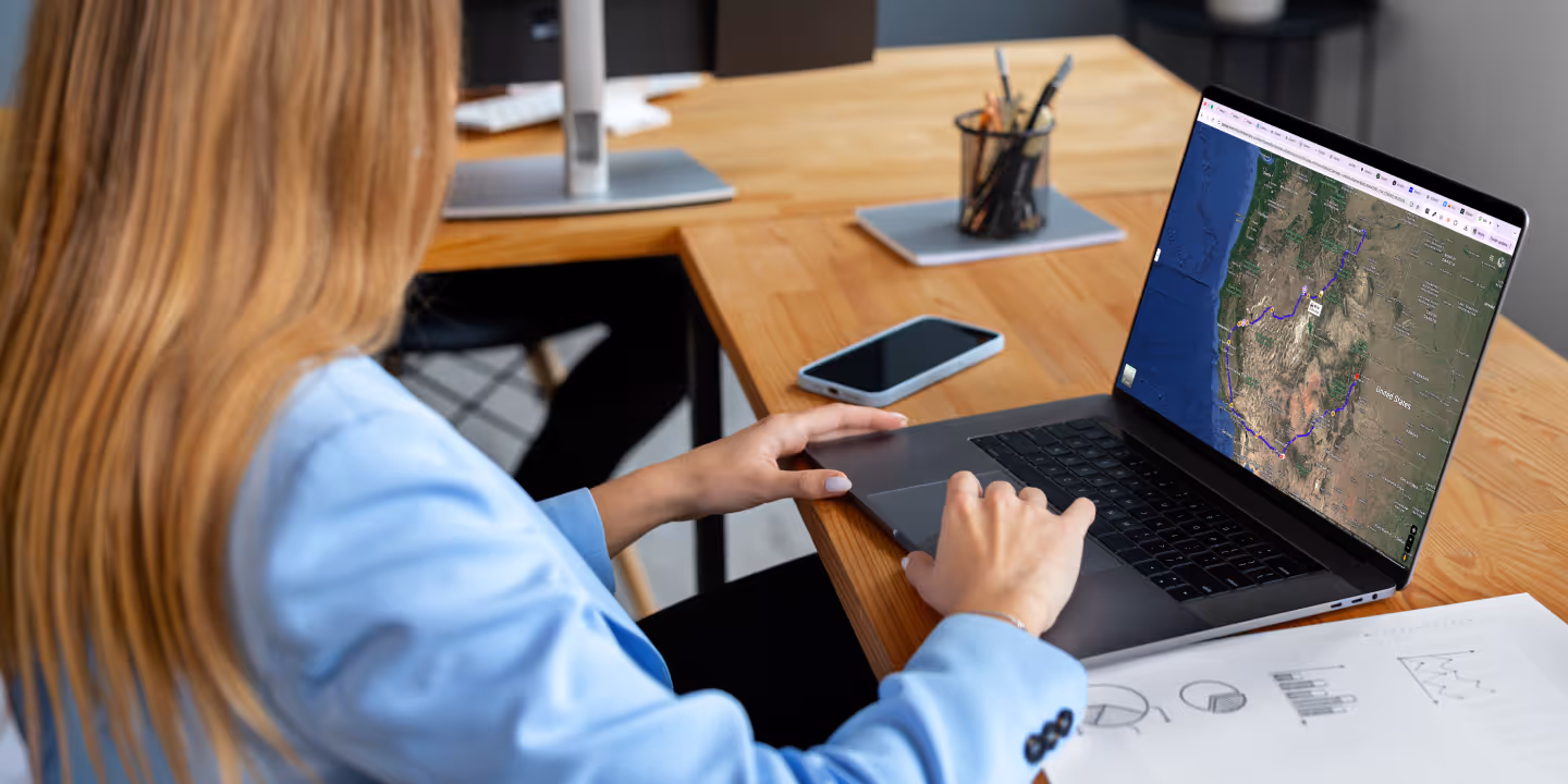 Person using a laptop showing a map with a marked route, sitting at a desk with a smartphone and documents with charts.